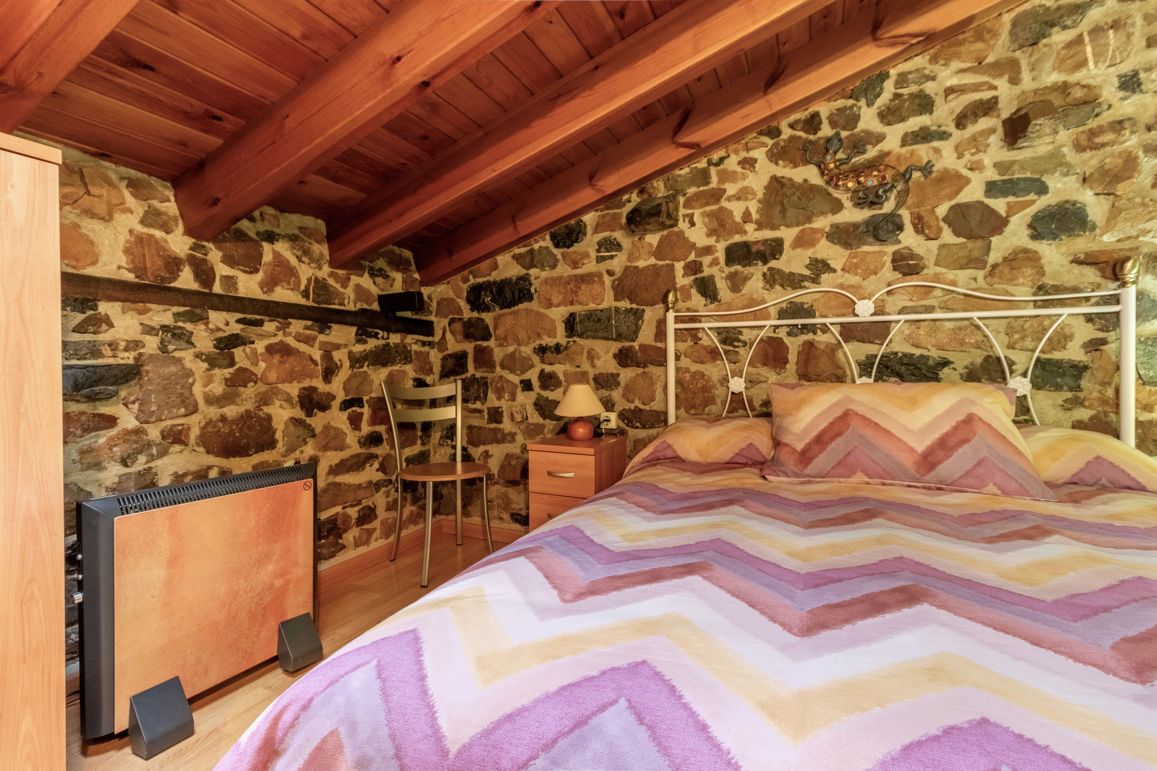 Een slaapkamer met een stenen muur, een bed met een deken met een zigzaggeluid, een houten plafond en een nachtkastje met een lamp.