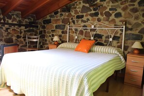 Quarto com parede de pedra, cama com roupa de cama listrada, travesseiro laranja, duas luminárias de cabeceira e mesa de cabeceira de madeira.