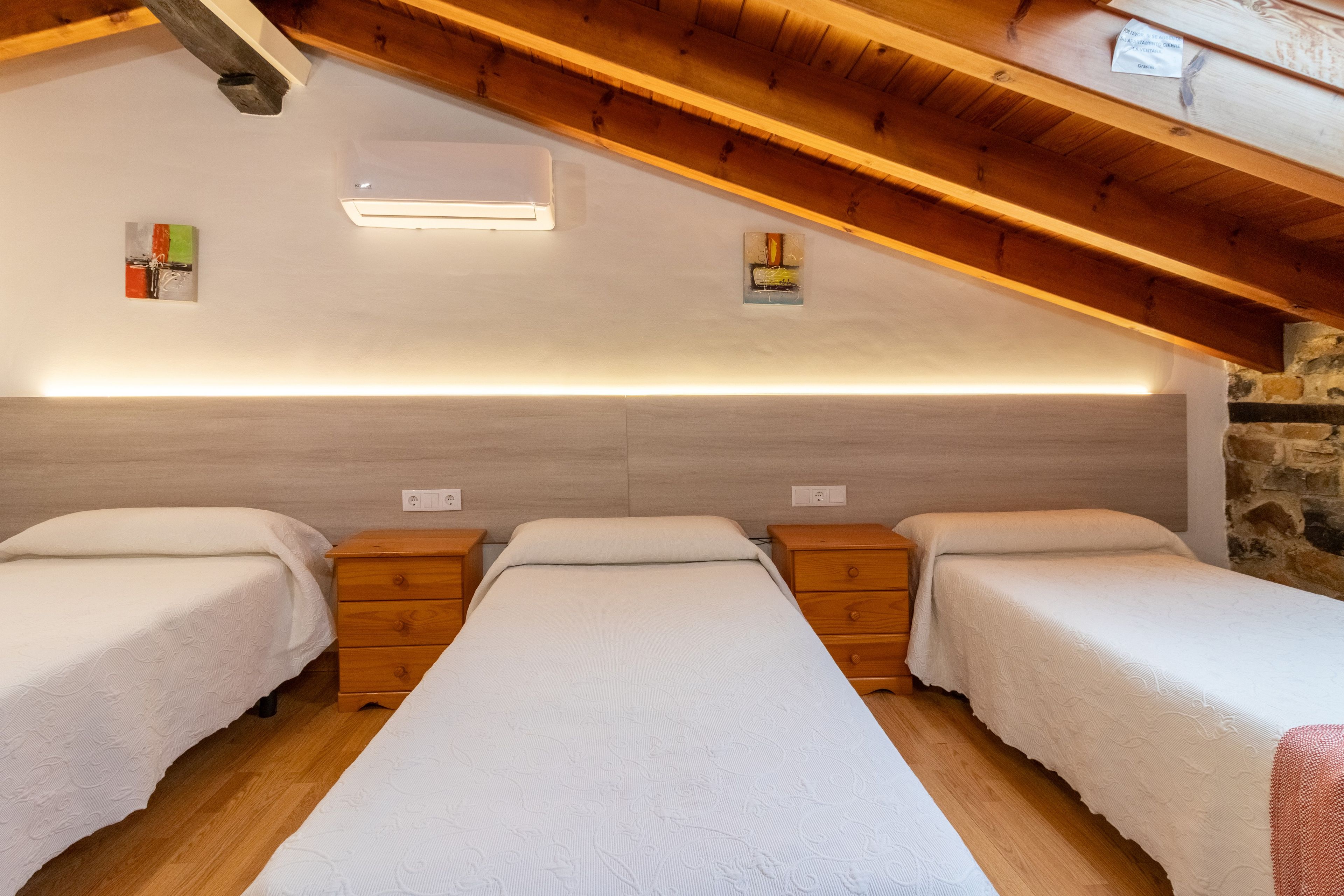 Une chambre avec trois lits simples alignés contre un mur, une tête de lit en bois et un plafond en pente avec des poutres apparentes.