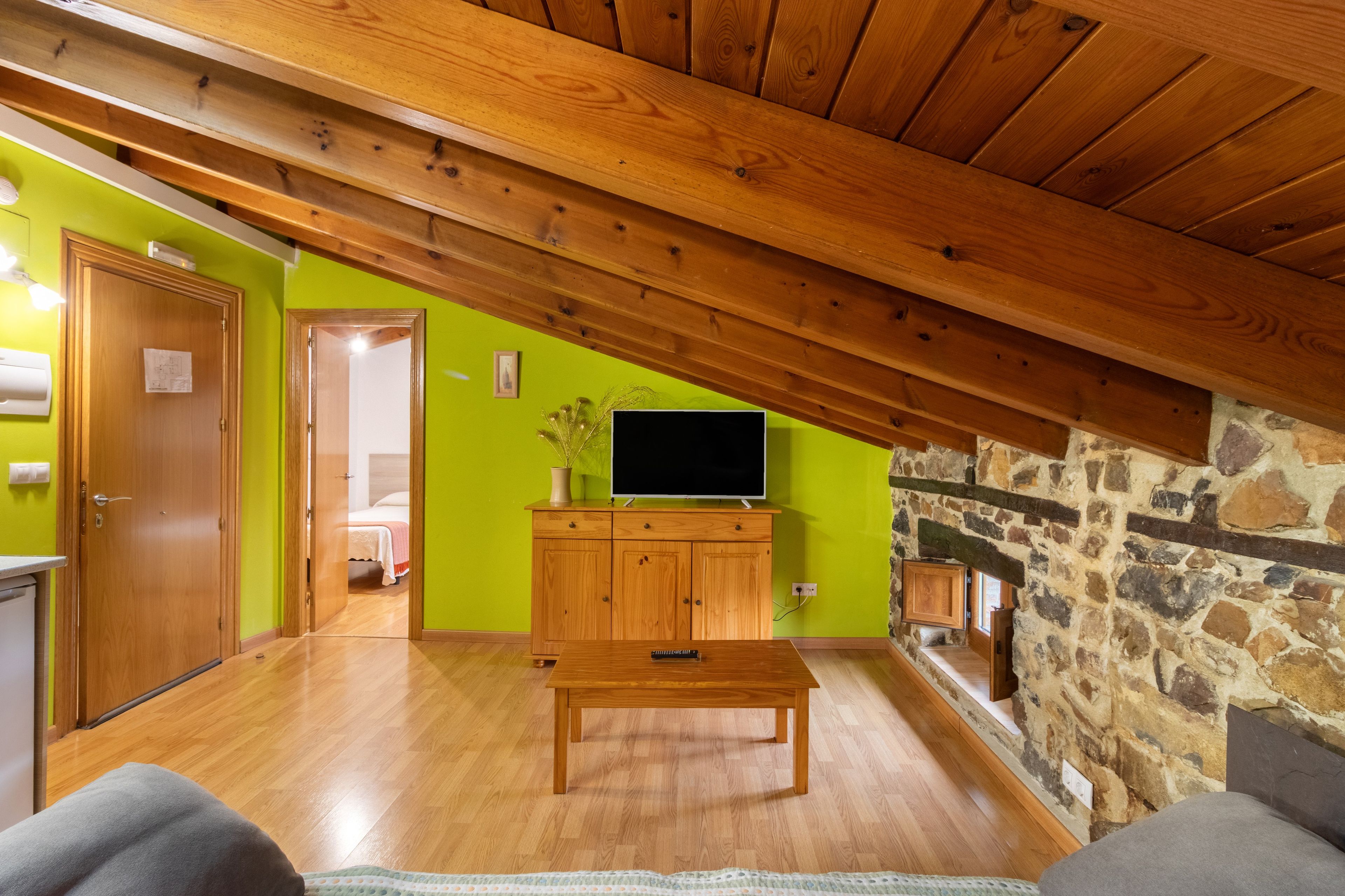 Una habitación con techo de madera, pared de piedra, televisor sobre un mueble de madera y una cama al fondo.