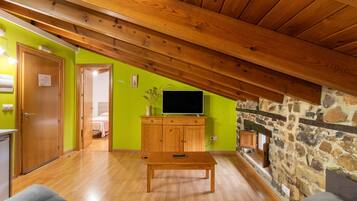 Una habitación con techo de madera, pared de piedra, televisor sobre un mueble de madera y una cama al fondo.