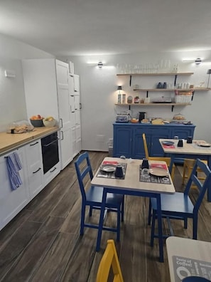 A compact kitchen with white cabinets, a blue dining table, and chairs. There are shelves above the counter with various items. - B&B Blue Dragonfly (Paesana)