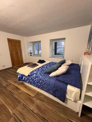 Un dormitorio con cama, dos ventanas, una puerta y piso de madera.