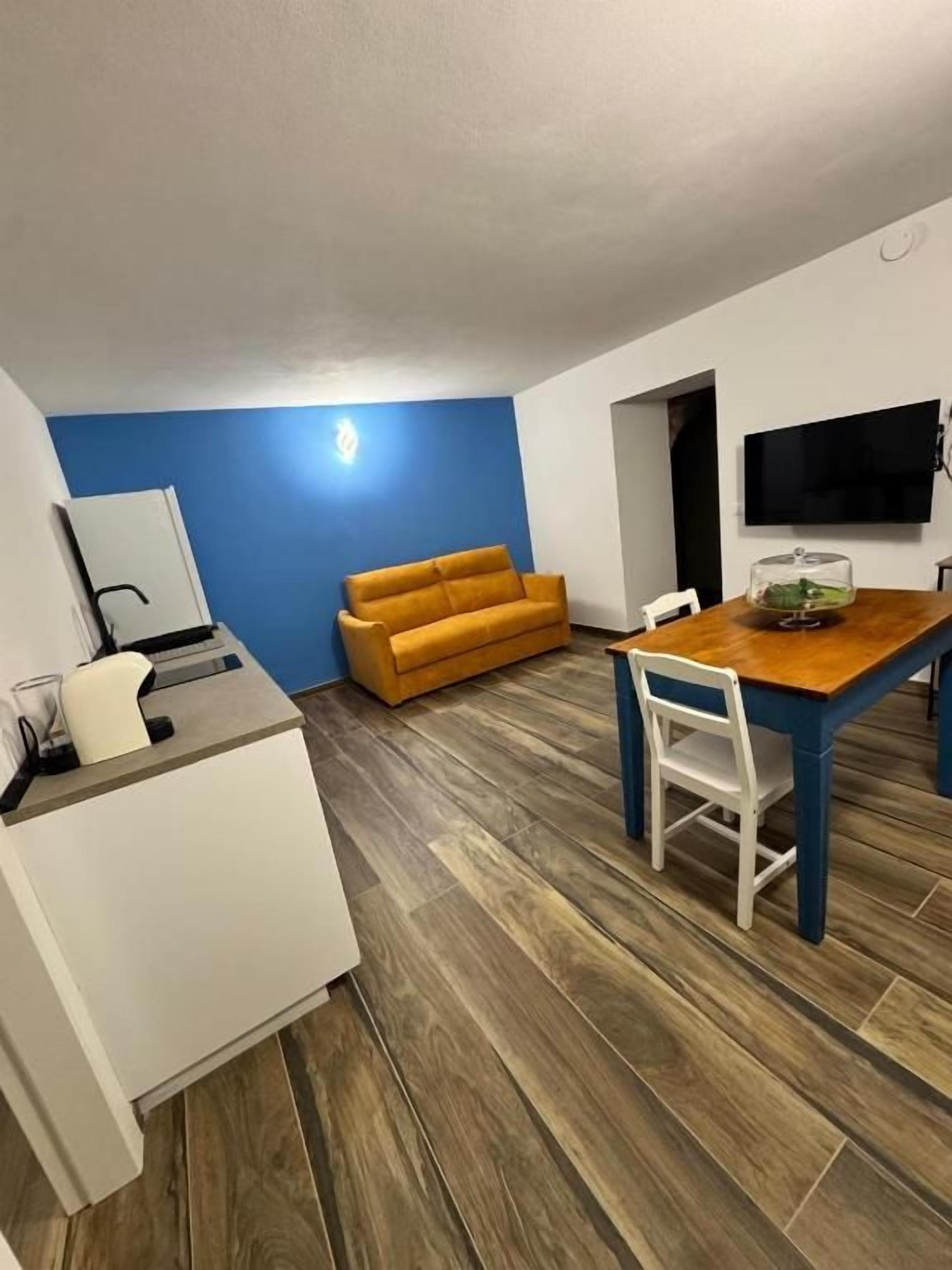 A compact living space with a kitchenette, a sofa, a dining table with chairs, and a television mounted on the wall.
