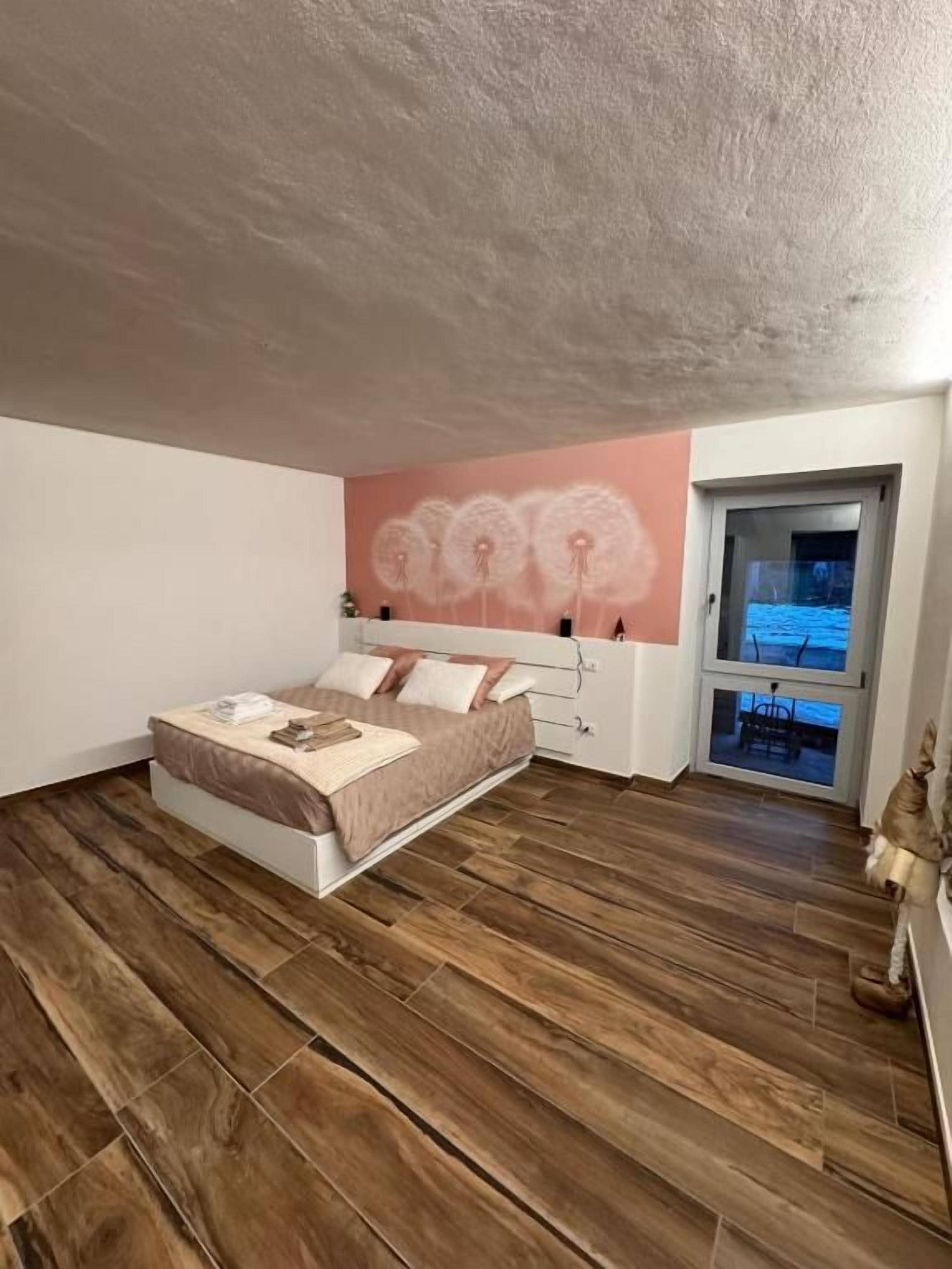 Ein Schlafzimmer mit einem Bett, einer rosa Wandmalerei, Holzboden und einer Tür, die auf einen Balkon führt.