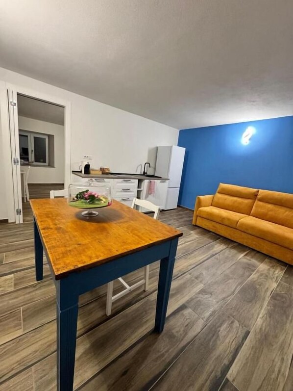 A room with a wooden dining table, a sofa, and a kitchenette in the background.