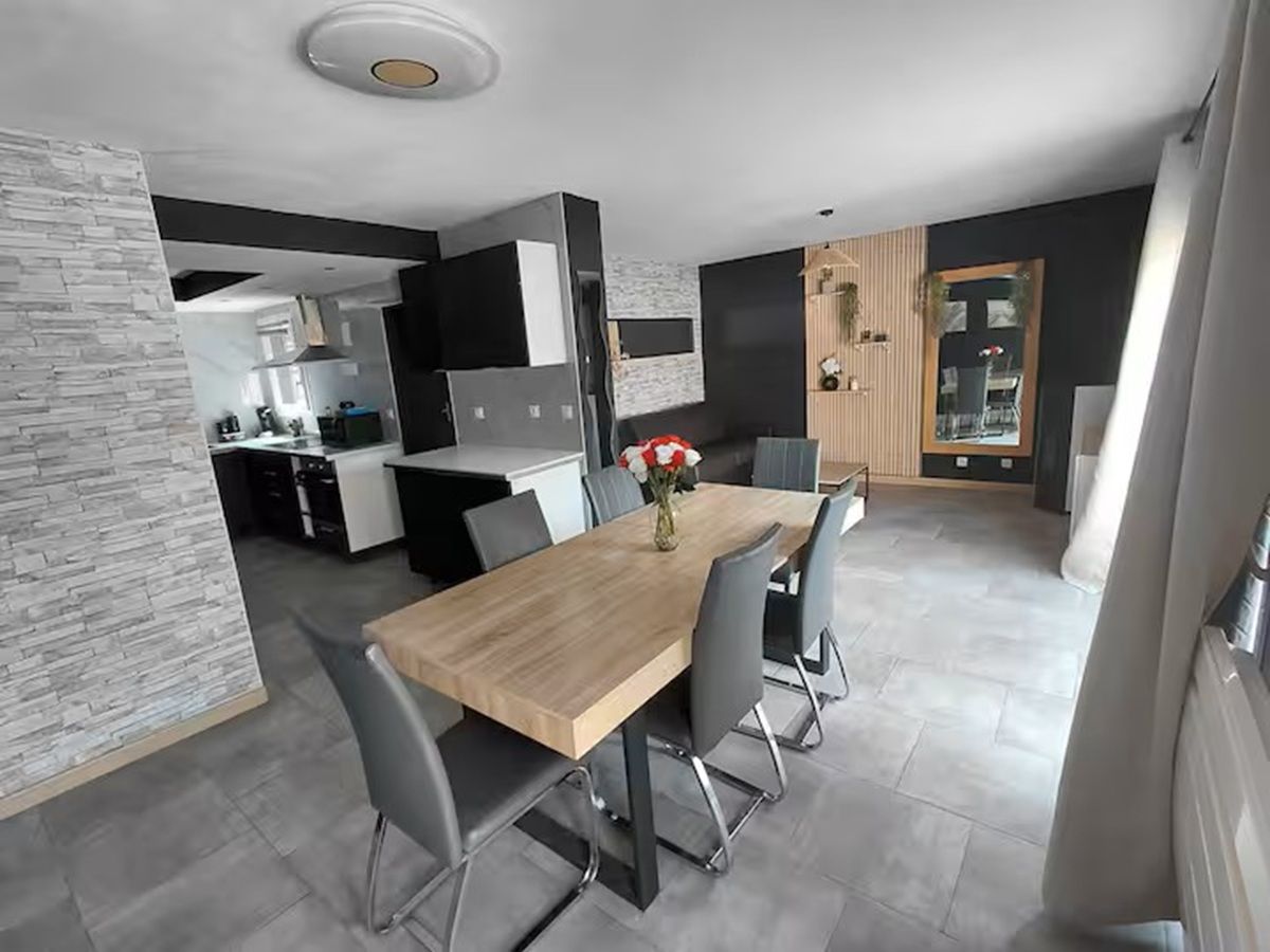 Un comedor moderno con una mesa de madera y sillas grises, una cocina al fondo y un jarrón decorativo con flores sobre la mesa.