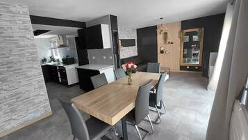 Un comedor moderno con una mesa de madera y sillas grises, una cocina al fondo y un jarrón decorativo con flores sobre la mesa.