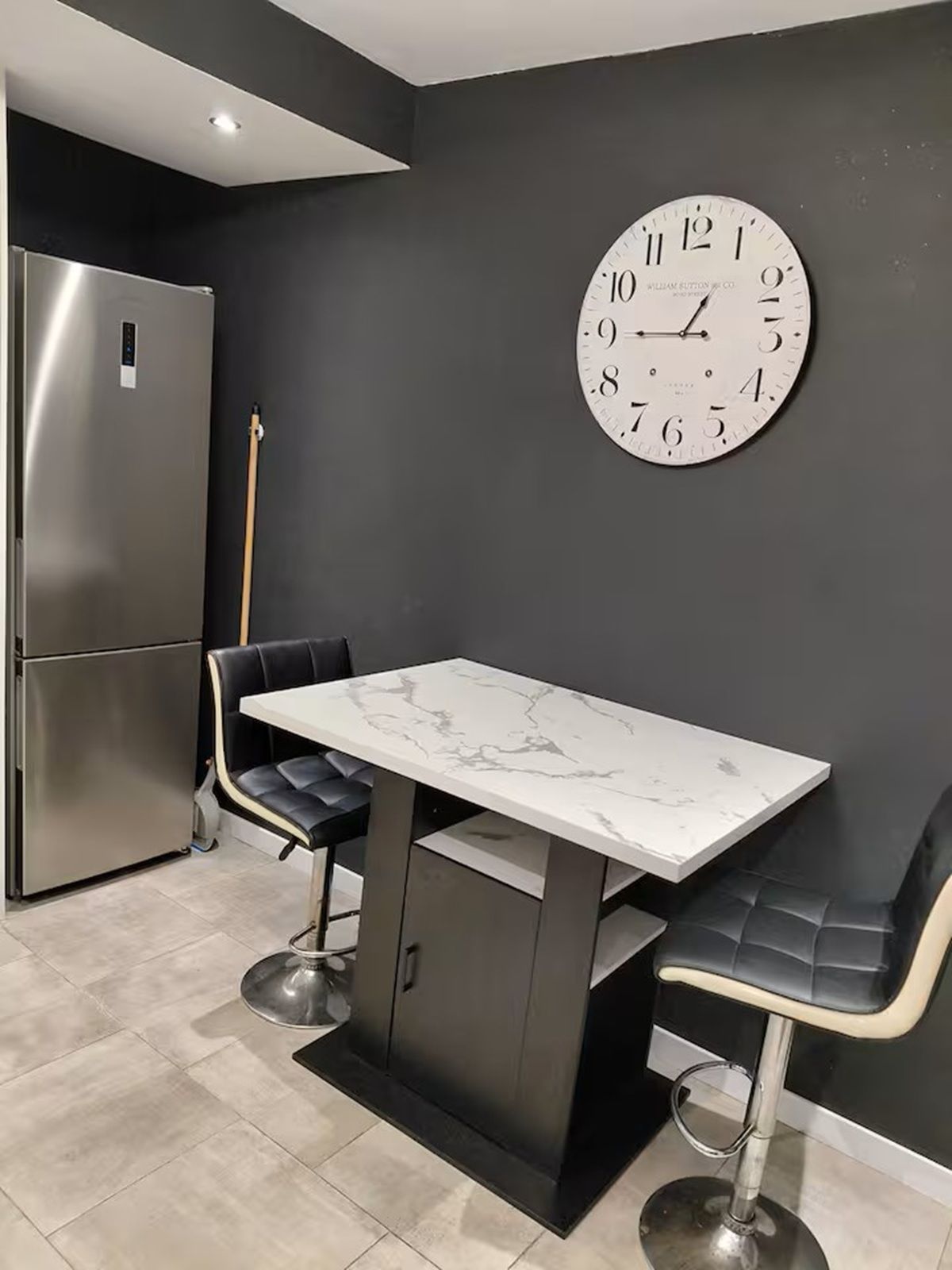 Eine moderne Küche mit einem Tisch aus Marmor, schwarzen Stühlen, einem Edelstahlkühlschrank und einer großen Wanduhr.