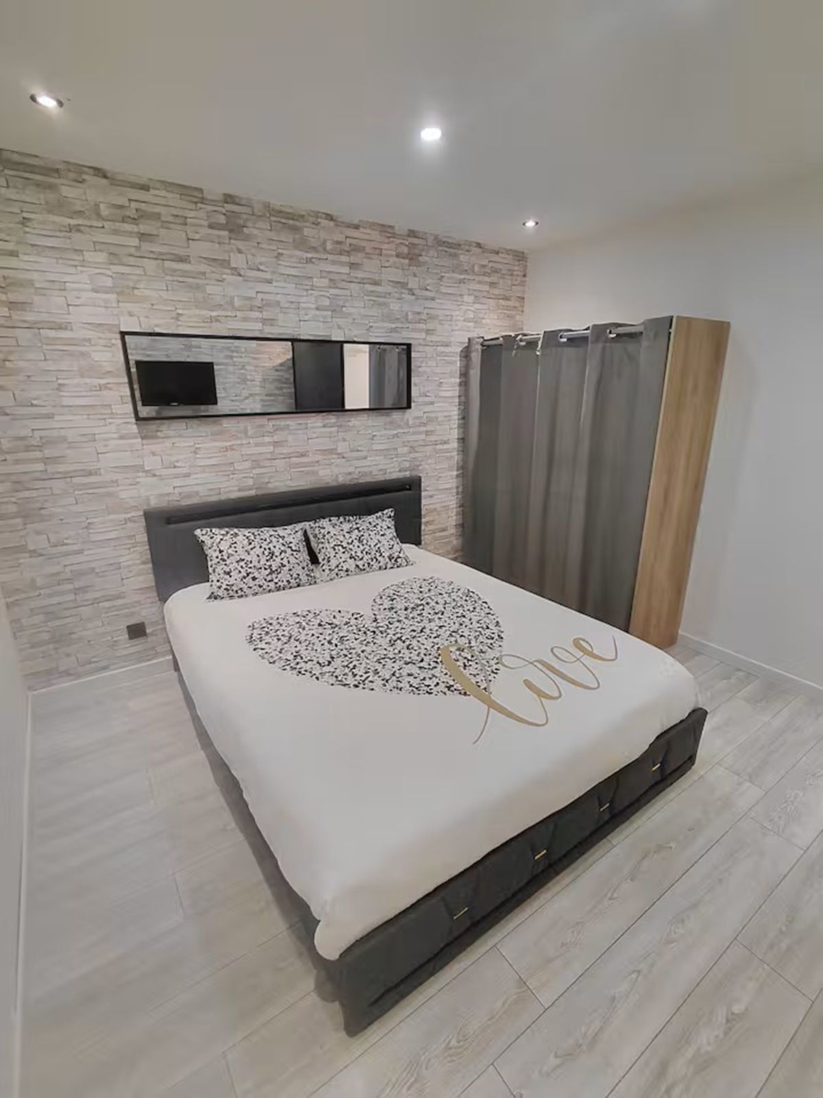 Un dormitorio moderno con pared de diseño de piedra, un televisor de pantalla plana, una cama con almohada en forma de corazón y una cortina gris.