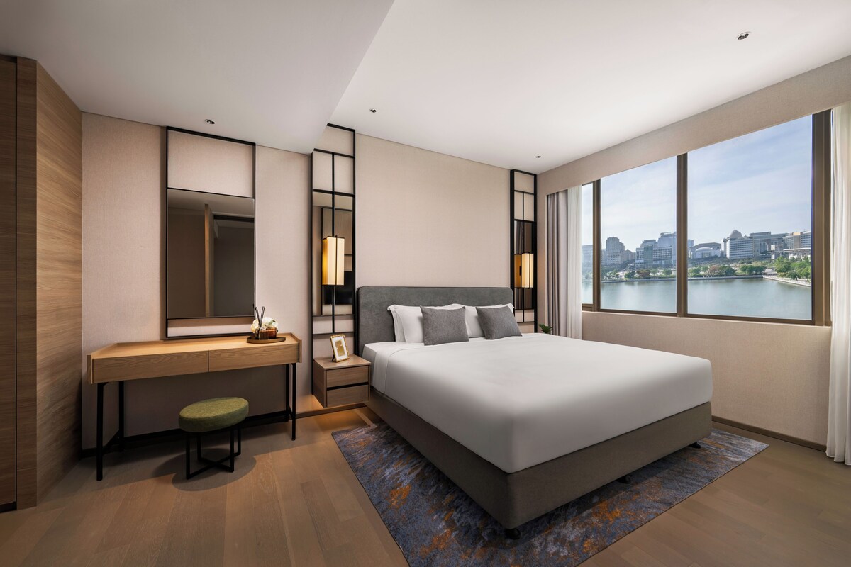 A modern hotel room with a large bed, a desk, a chair, a nightstand, a mirror, and a view of a cityscape.