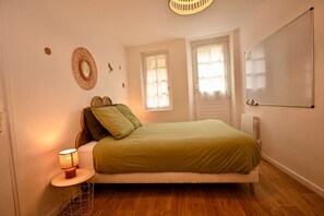 Un dormitori amb llit, una làmpada de nit, un mirall fixat a la paret i una finestra amb cortines.