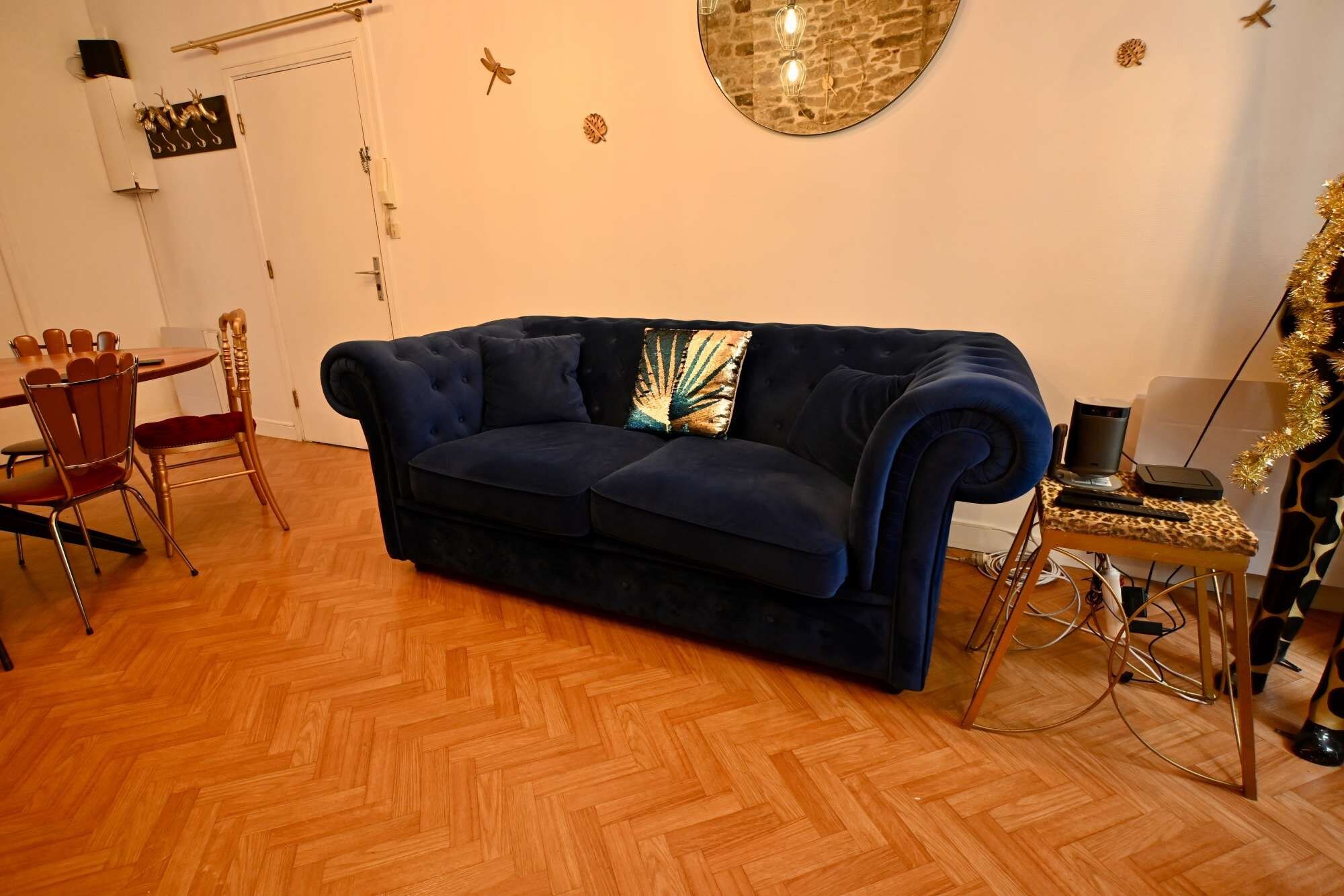 A room with a dark blue sofa, a wooden herringbone floor, a dining table with chairs, and a decorative mirror on the wall.