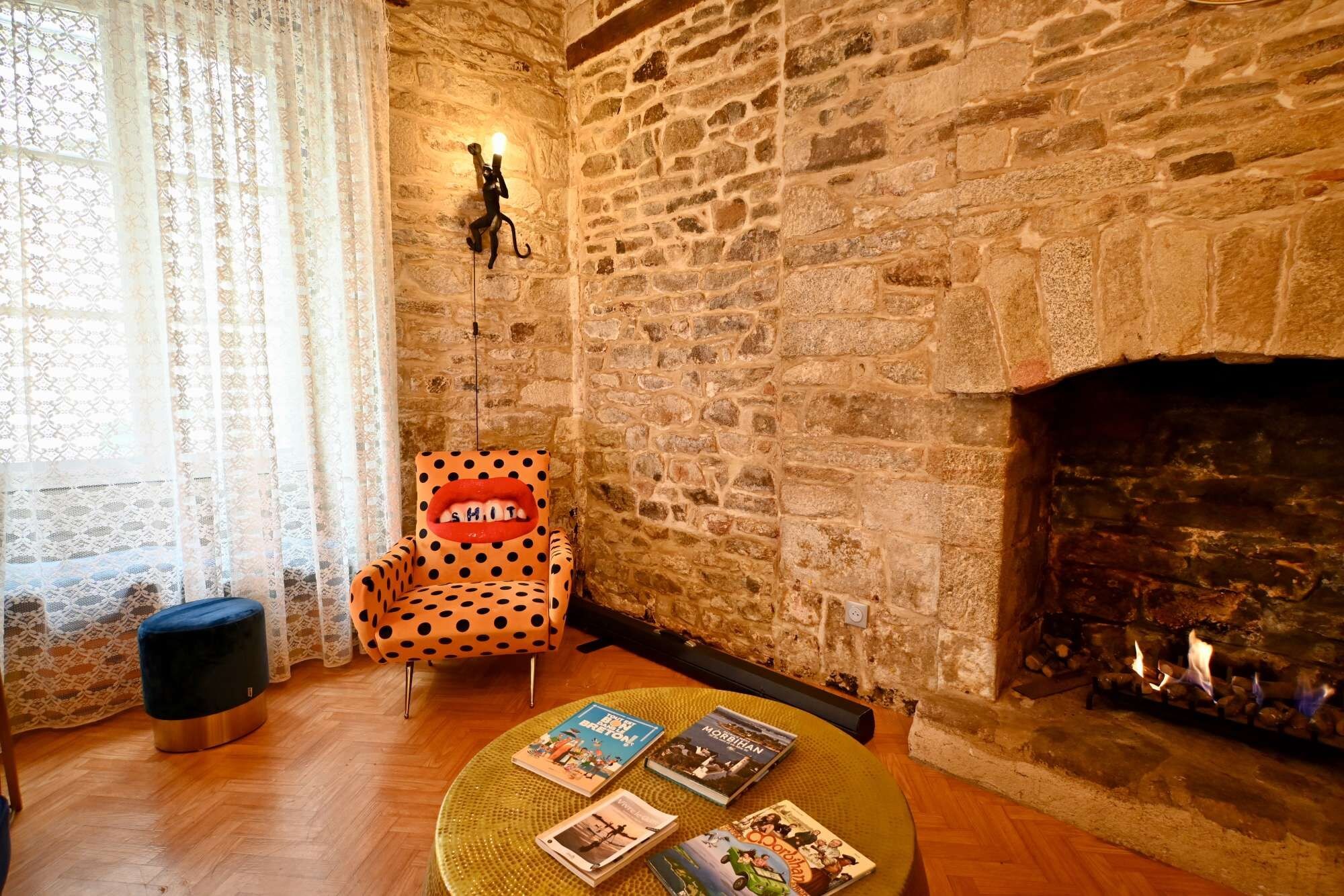 Quarto aconchegante com parede de pedra, lareira, cadeira com estampa de bolinhas e mesa redonda com livros.