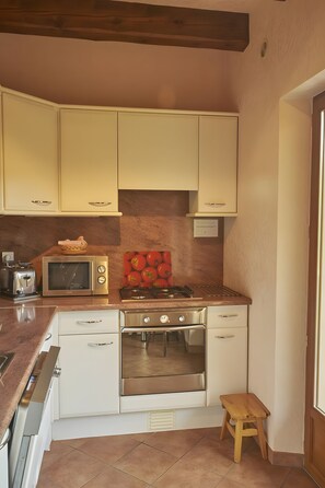 Een kleine keuken met witte kastjes, een magnetron, een gasfornuis en een klein houten krukje.