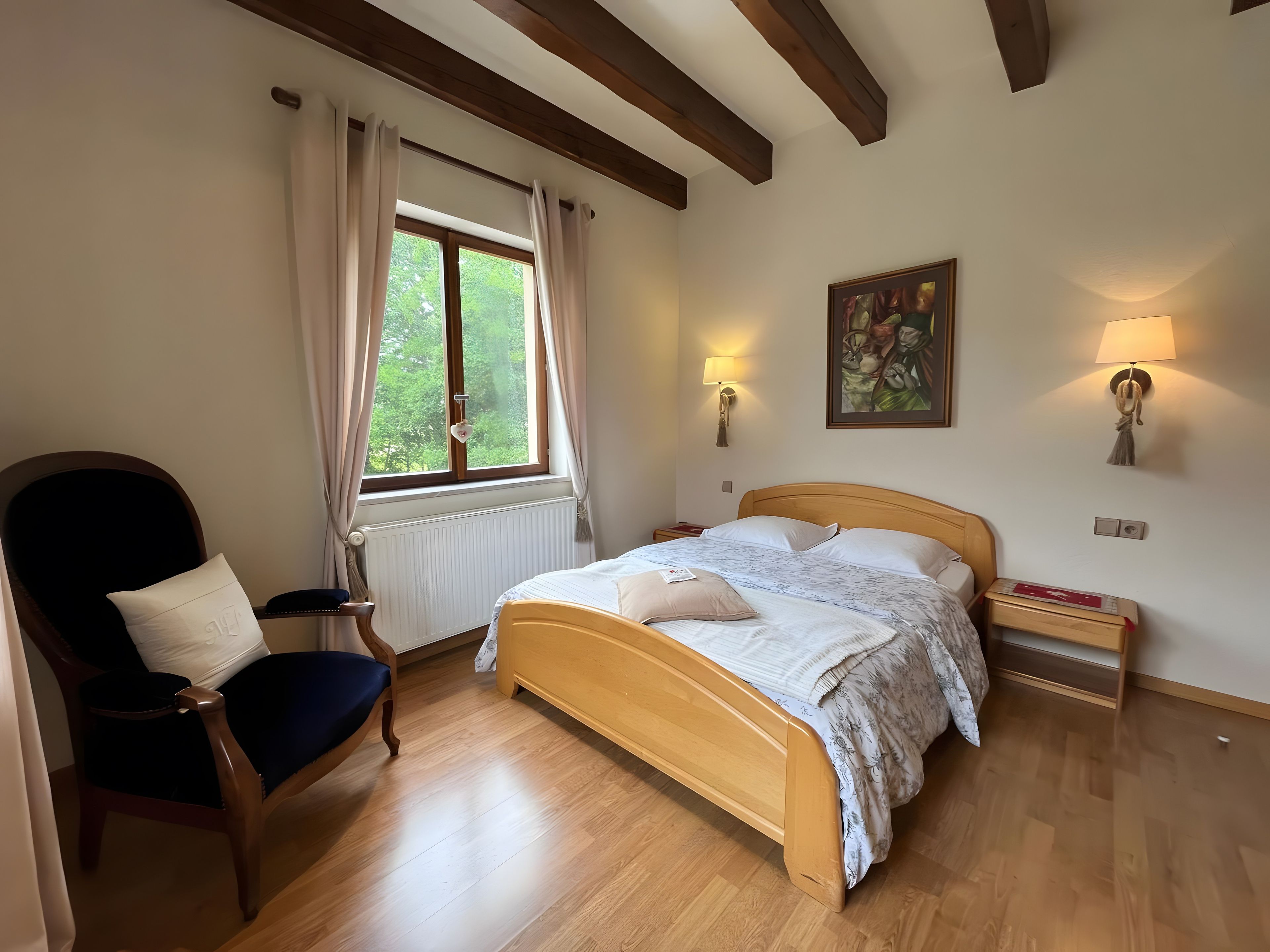 Ein Schlafzimmer mit einem hölzernen Bett, einem Sessel, einem Fenster mit Vorhängen, einem Heizkörper und Wandlampen.