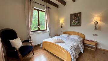 Ein Schlafzimmer mit einem hölzernen Bett, einem Sessel, einem Fenster mit Vorhängen, einem Heizkörper und Wandlampen.