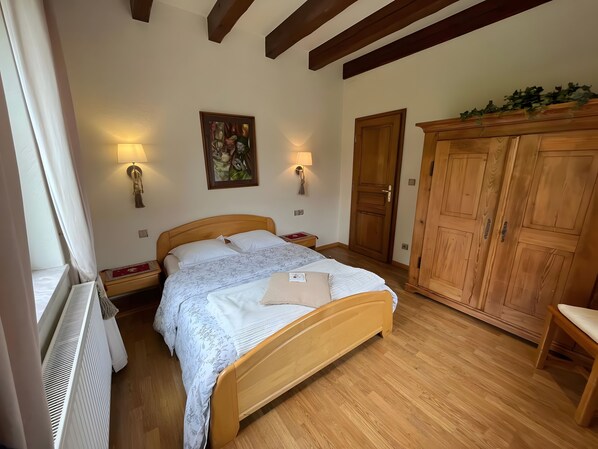 A bedroom with a wooden bed, two bedside lamps, a radiator, a wooden wardrobe, and a painting on the wall.