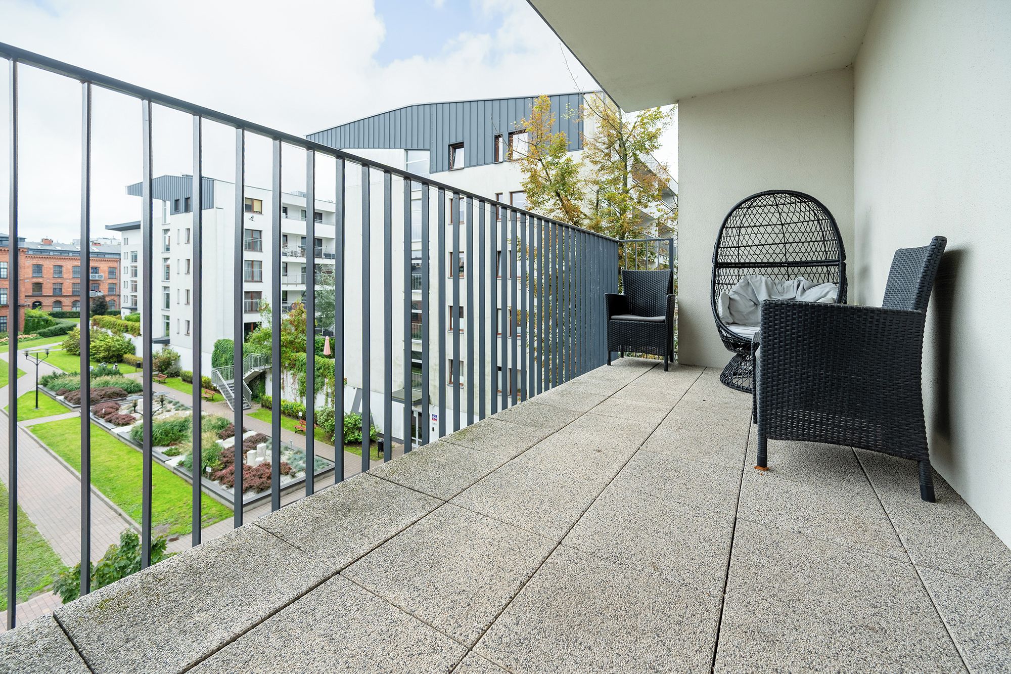 Un balcone con ringhiera in metallo, una sedia in vimini e vista su edifici e vegetazione.
