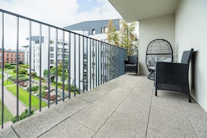 Un balcón con barandilla de metal, una silla de mimbre y vistas a edificios y zonas verdes.