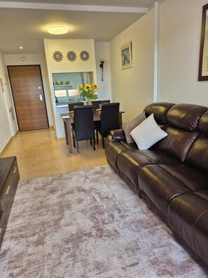 A living room with a brown leather sofa, a dining table with chairs, and a kitchen in the background. - Home away from Home in the Sun (MC)