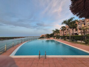 Ein Swimmingpool mit freiem Blick auf das Meer, umgeben von einem Zaun und Palmen, mit einem Gebäude im Hintergrund.