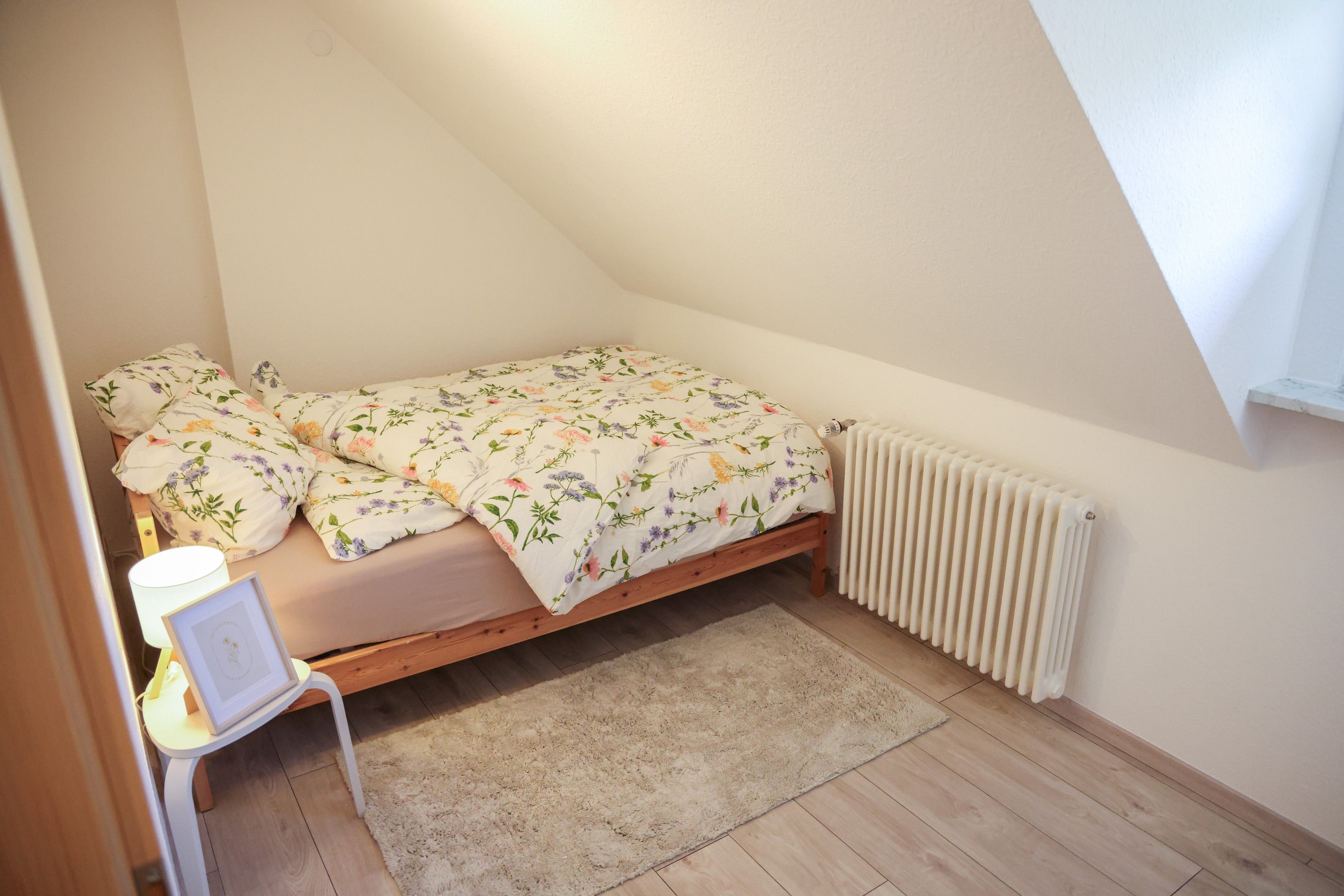 A small, single-bed room with a wooden frame, a white radiator, a white chair, and a fluffy rug.