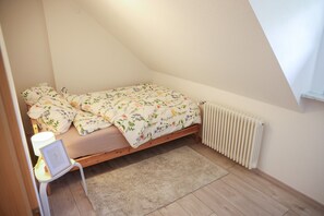 Une petite chambre avec un lit simple, un cadre en bois, un radiateur blanc, une chaise blanche et un tapis moelleux.