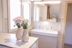 Un baño con un lavamanos blanco, espejo y dos jarrones blancos con flores sobre una encimera.