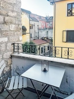 Un balcón con una mesa y sillas blancas, barandilla metálica y vista a edificios coloridos al fondo.