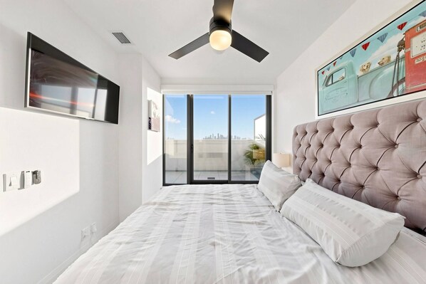 Un dormitorio con una cama grande, un ventilador de techo, vistas a la ciudad a través de la ventana y un cuadro enmarcado en la pared.