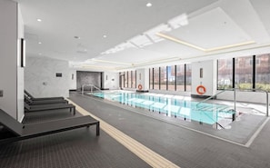 Una piscina interior moderna con paredes de vidrio transparente, suelo de baldosas y una zona de bancos.