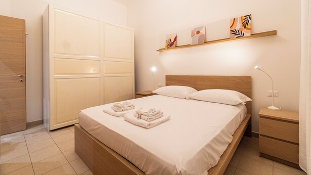 A neatly made bed with towels, a wooden wardrobe, a bedside table with a lamp, and artwork on the wall. - Achillini 9 (Bologna)