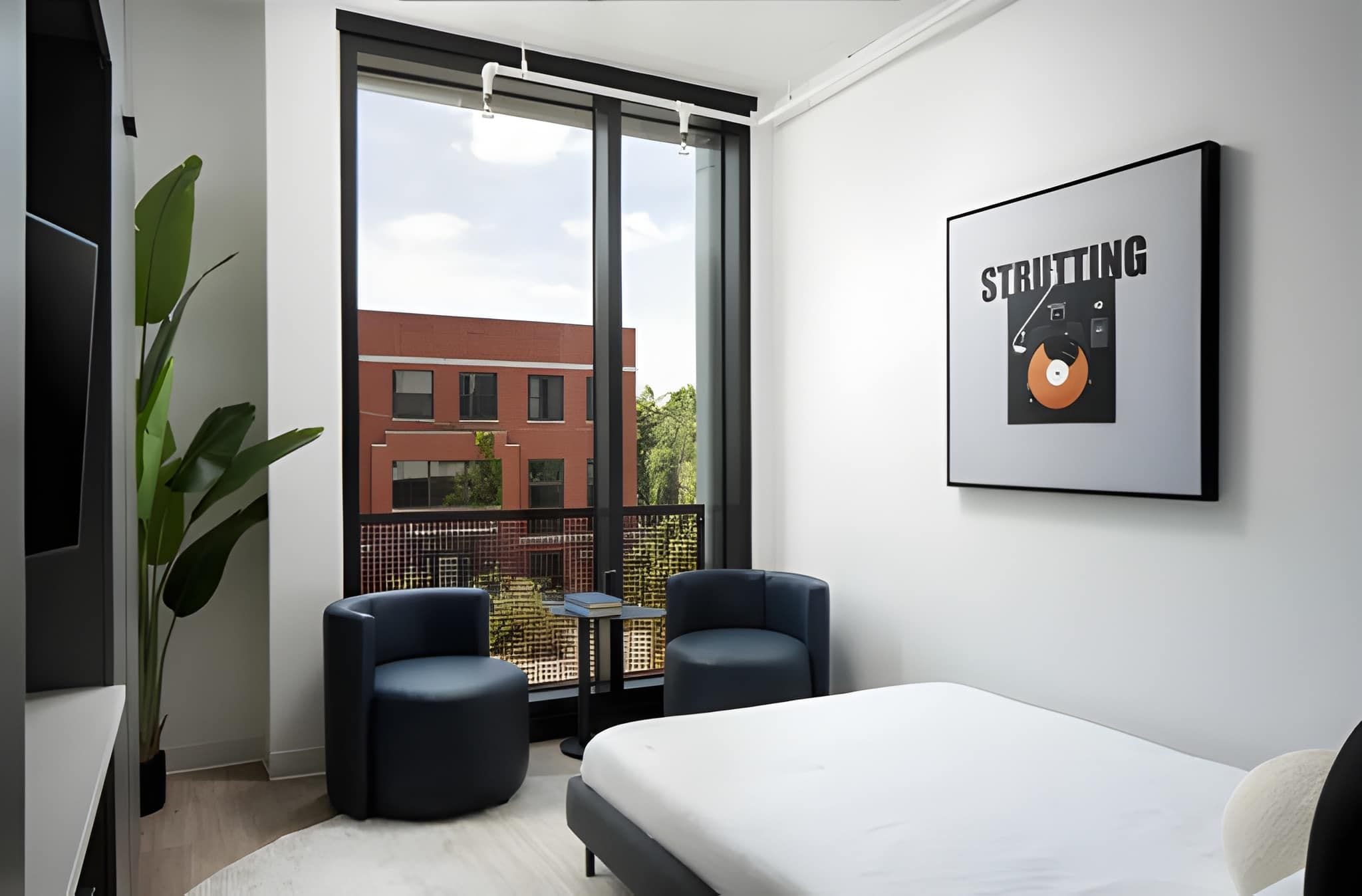 Une chambre d’hôtel moderne avec une grande fenêtre, deux fauteuils, un lit et un tableau encadré au mur.