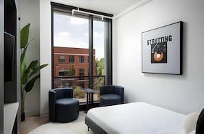 A modern hotel room with a large window, two chairs, a bed, and a framed picture on the wall.