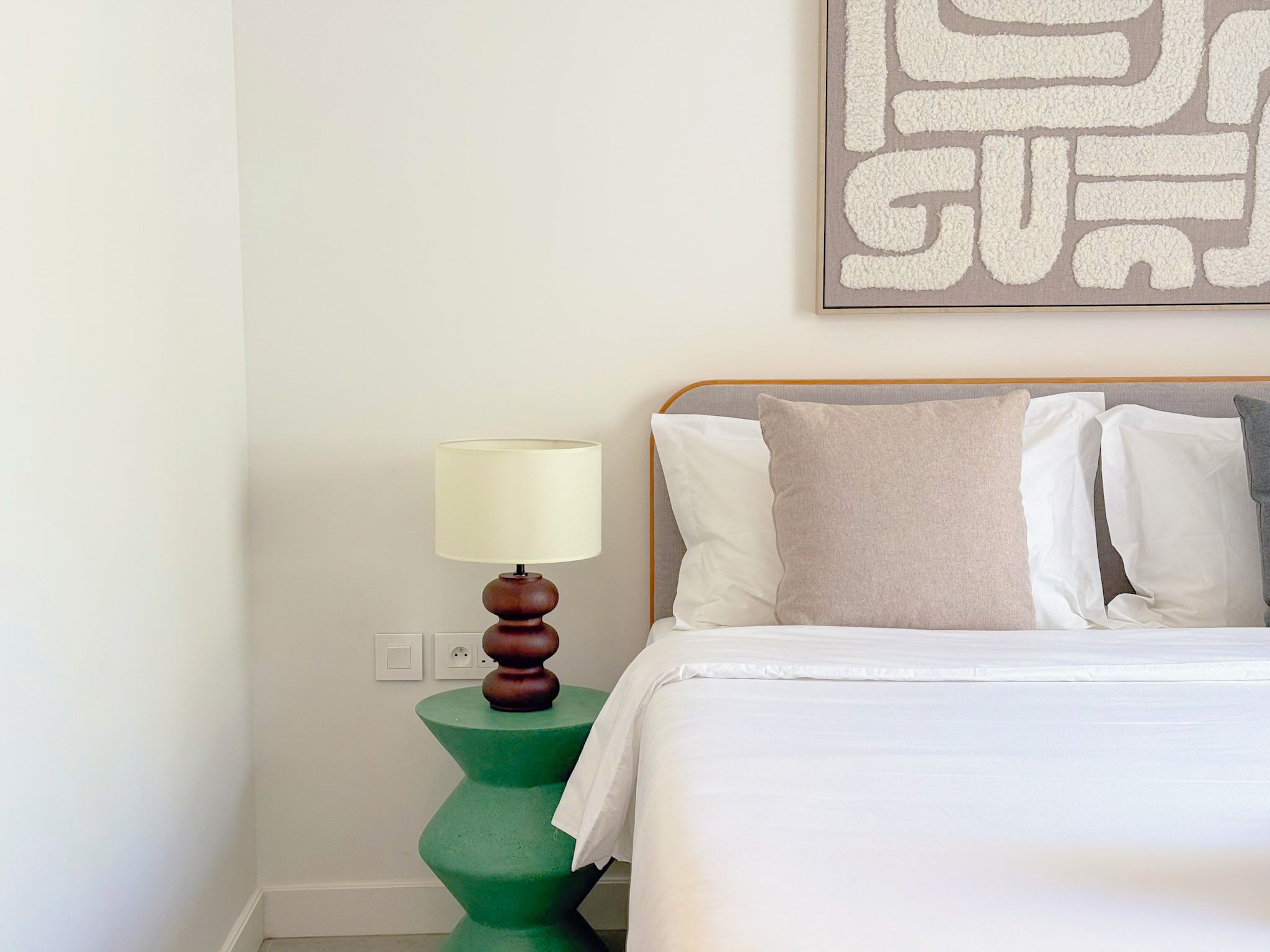A neatly made bed with white linens, a green nightstand with a lamp, and a wall-mounted artwork.