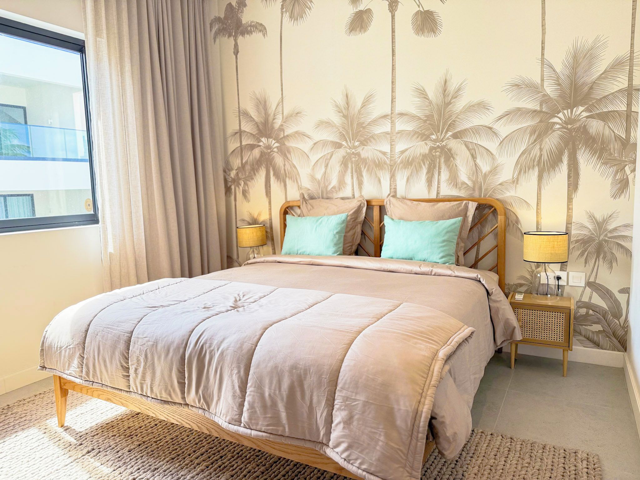 A bedroom with a bed, curtains, a wall mural, a nightstand, and a lamp.