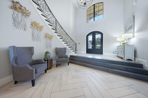 Um hall espaçoso com piso em padrão herringbone, uma escadaria imponente com grades de metal ornamentadas e uma ampla porta em arco.