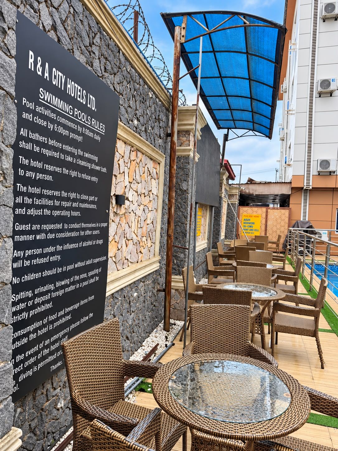 A poolside area with wicker furniture and a glass table, a sign detailing pool rules, and a stone wall.