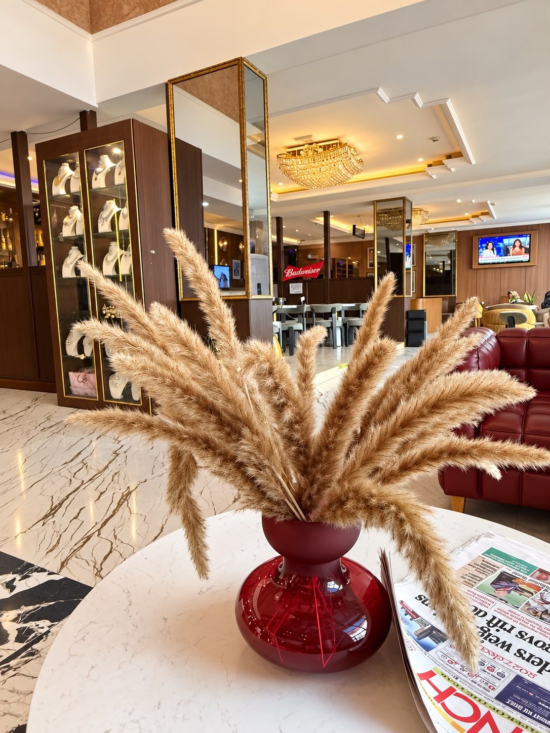 Un moderno vestíbulo de hotel con una mesa redonda de mármol, un jarrón rojo con plumas de pampa, un sofá de cuero rojo y un letrero de Budweiser.