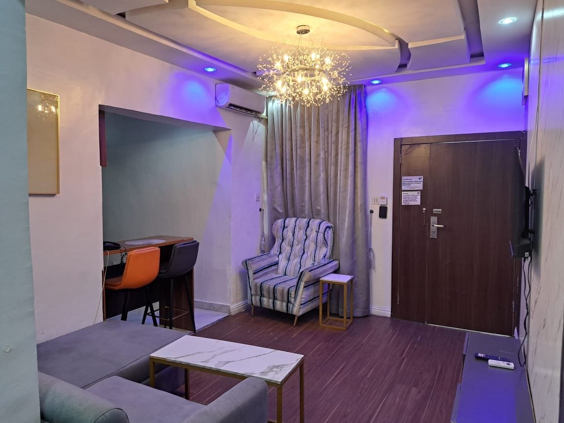 A modern room with a sofa, a chair, a small table, a desk with chairs, a chandelier, and a door.