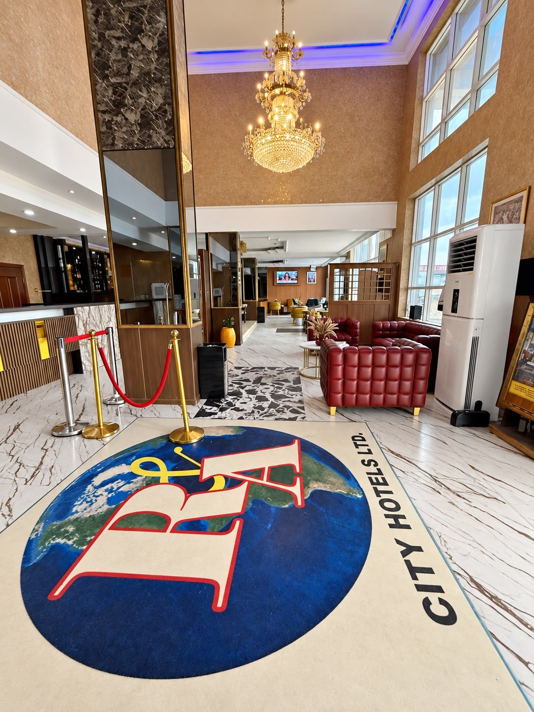 A hotel lobby with a large chandelier, a reception desk, a red leather sofa, and a floor design with a globe and the hotel's name.