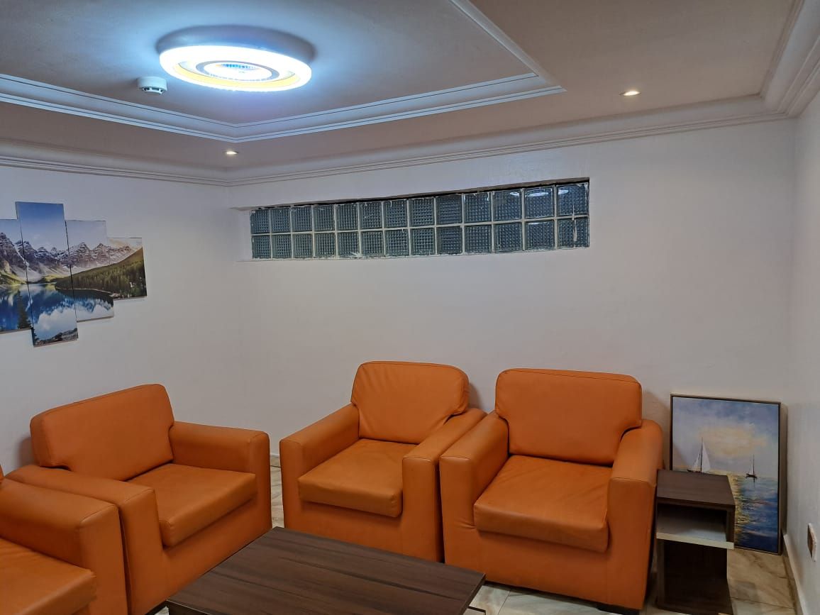 Una habitación con tres sillas naranjas, una mesa de centro de madera, una lámpara de techo, un ventanal con persianas y un cuadro en la pared.