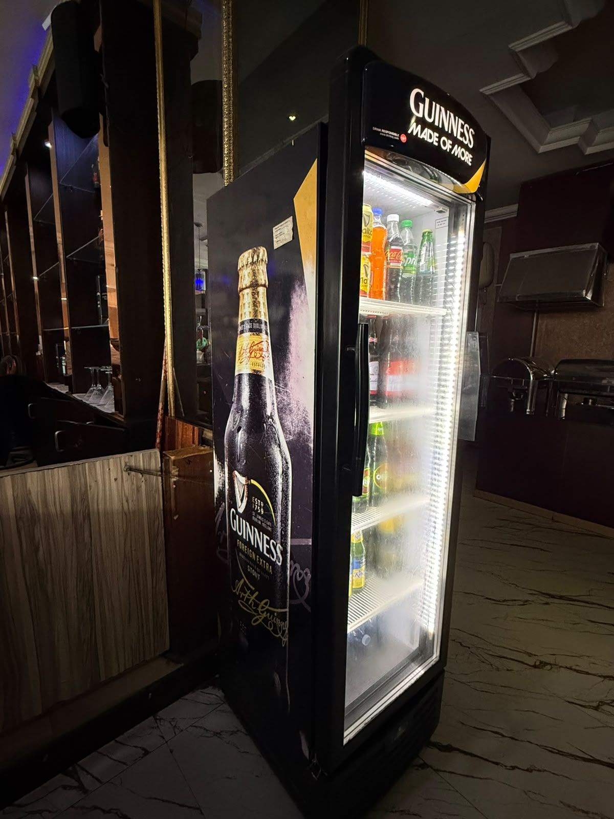 Een koelkast gevuld met drankjes, waaronder Guinness bier, in een bar met een houten toonbank en een spiegel.