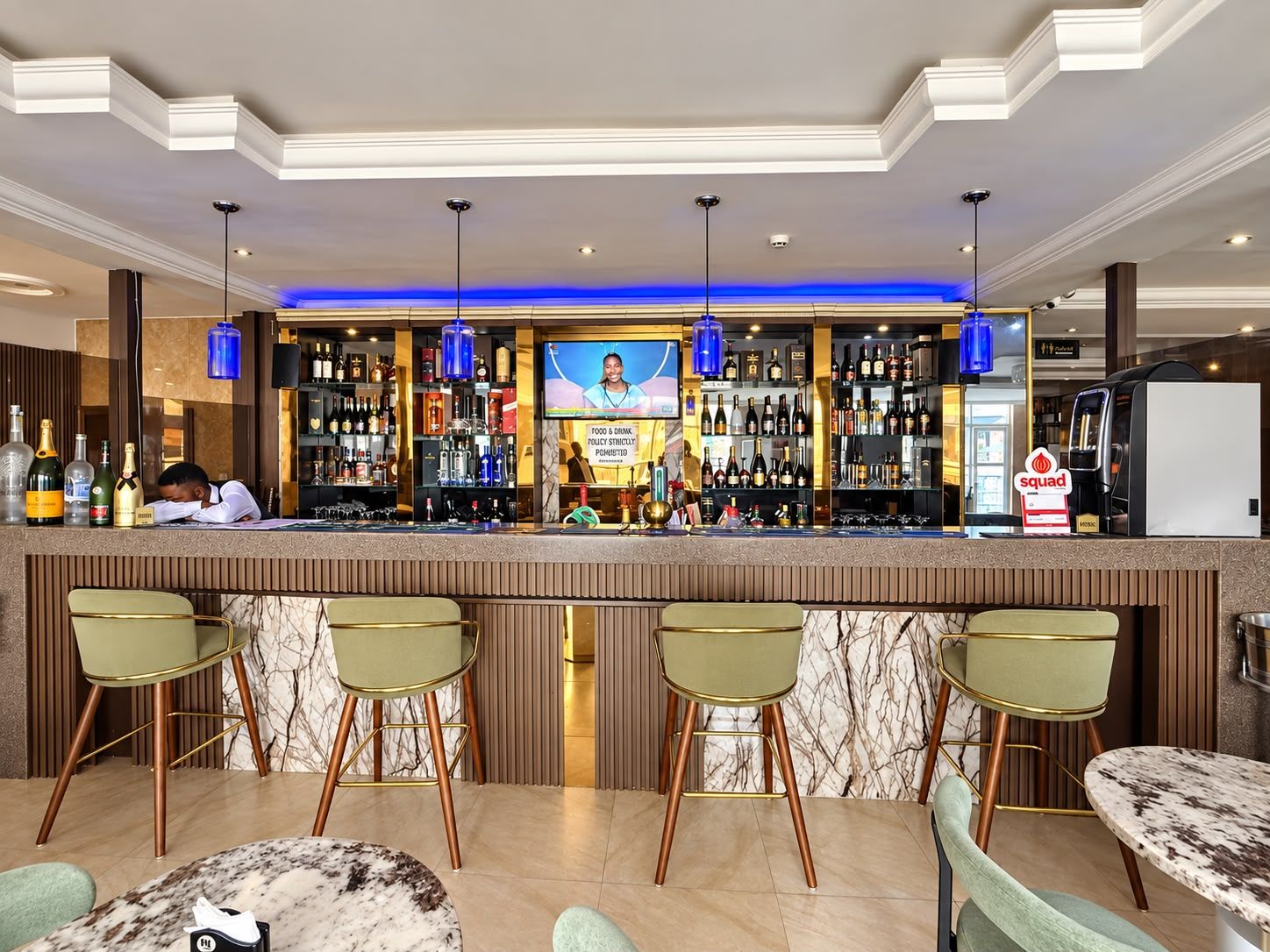A bar area with a counter, multiple bar stools, and a television screen displaying a person.