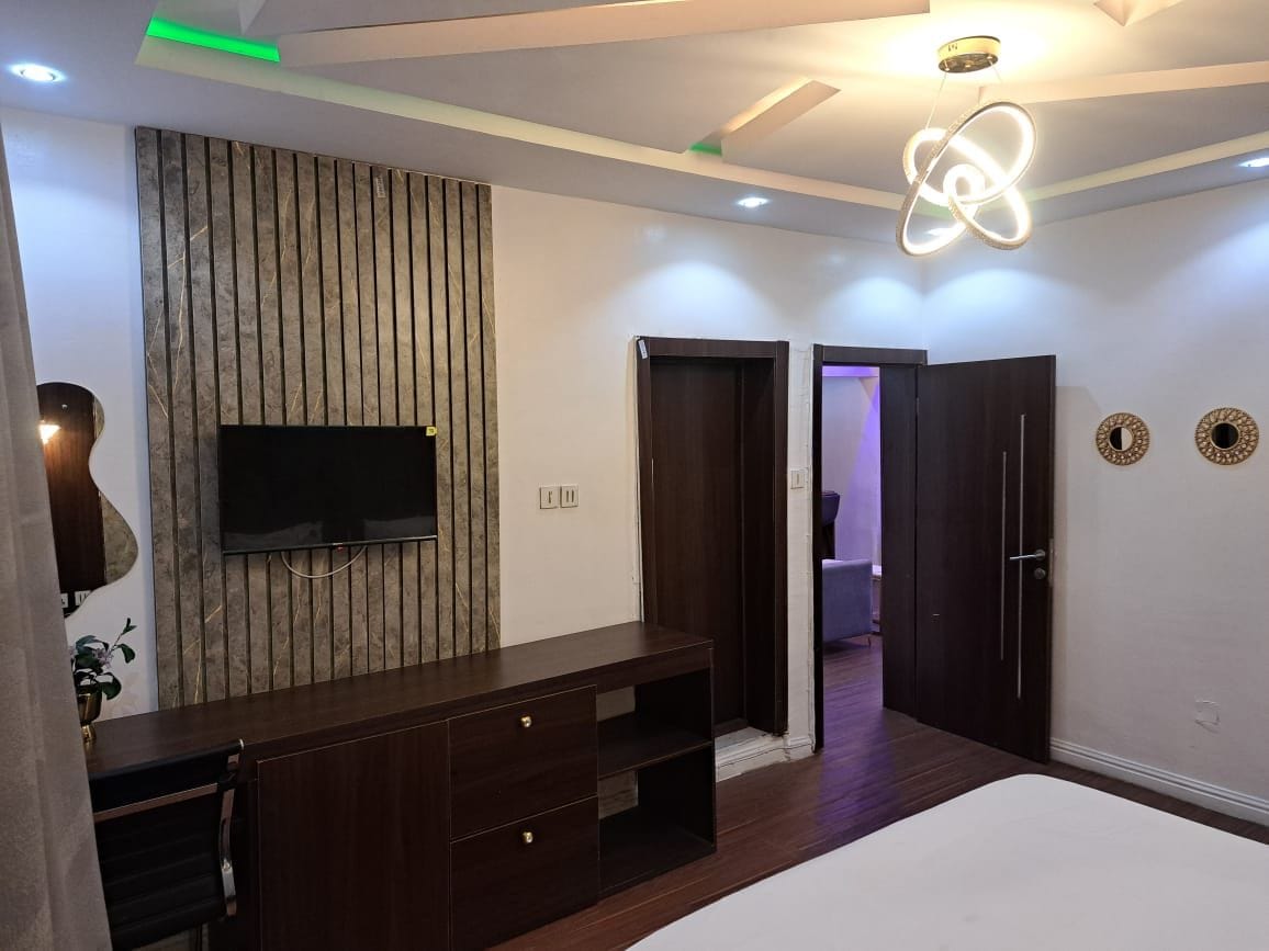 Un dormitorio moderno con una cama grande, un televisor montado en una pared de paneles de madera, un aparador de madera oscura y una puerta que da a otra habitación.