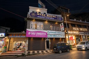 Un edificio con un cartel que dice 'Stay Royal Homestay / Guest House' y tiendas adyacentes, incluyendo 'Baru Sweets' y 'Bawa Sweets'.