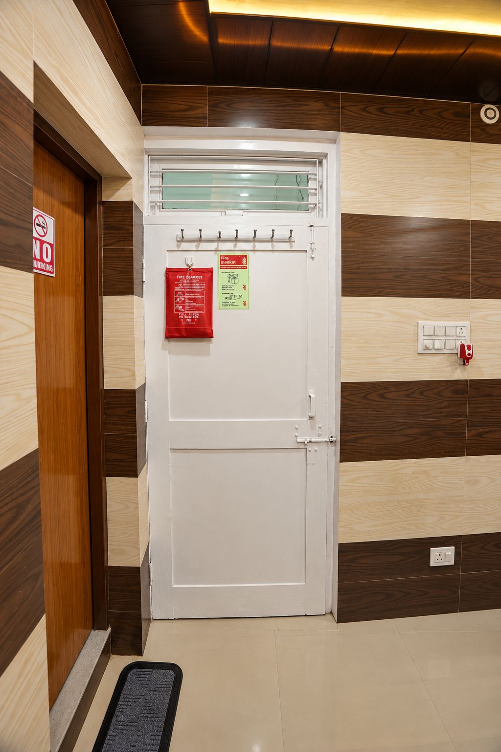 A hotel room entrance with a white door, a fire blanket, and a 'No Smoking' sign.