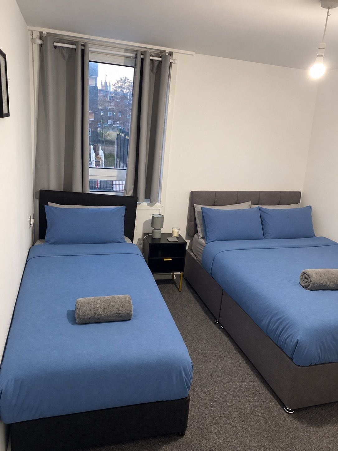 Habitación de hotel con dos camas, una mesita de noche, una lámpara y vista a la ciudad.