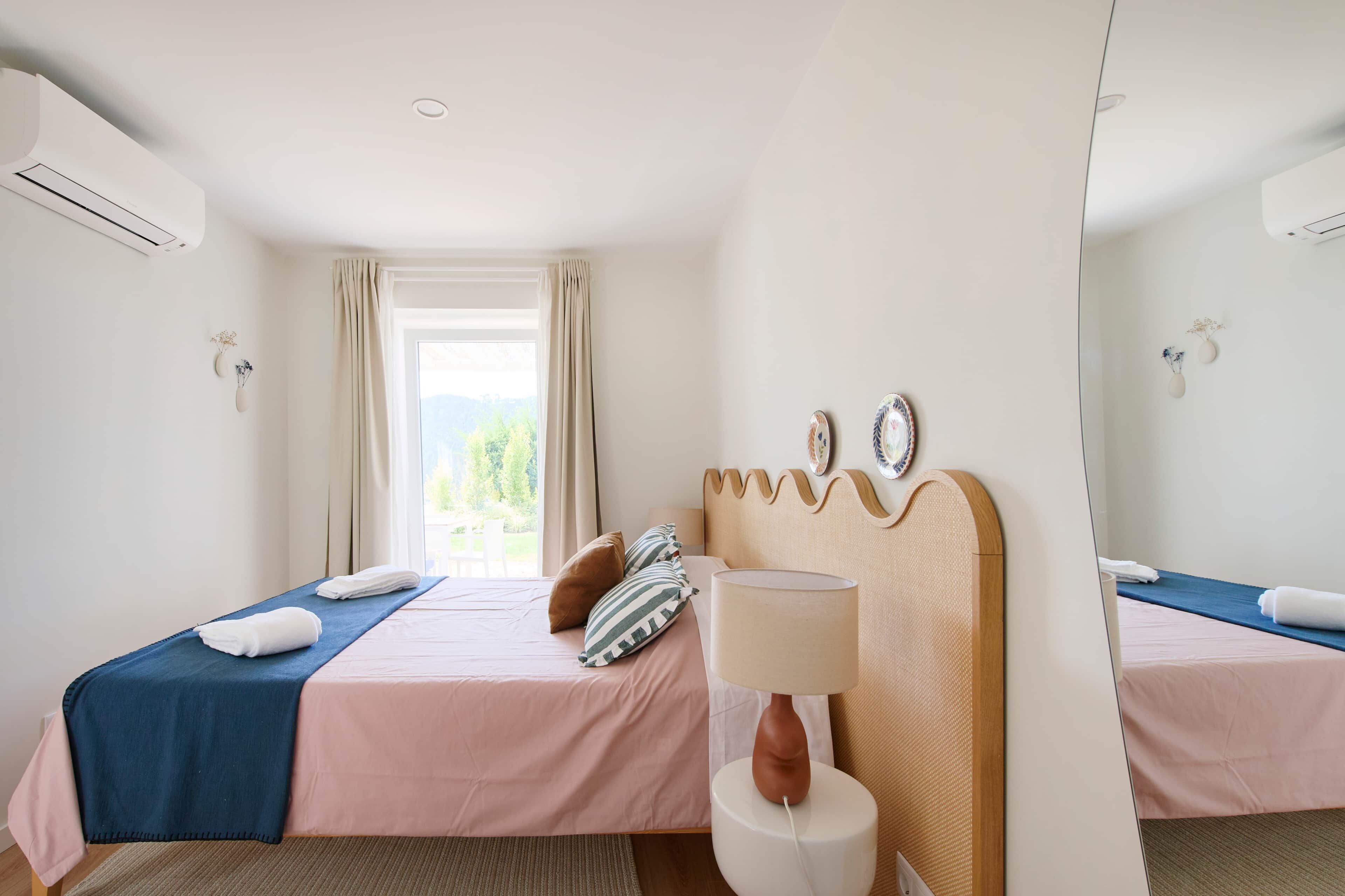 A hotel room with two beds, a window with curtains, a bedside table with a lamp, and a wall-mounted air conditioner.